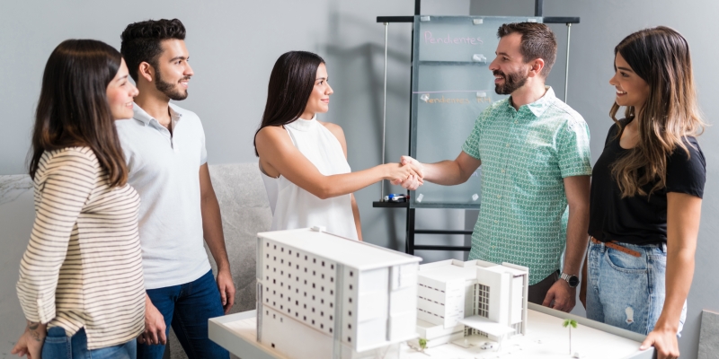 Business partners shaking hands with engineer by architectural model in meeting
